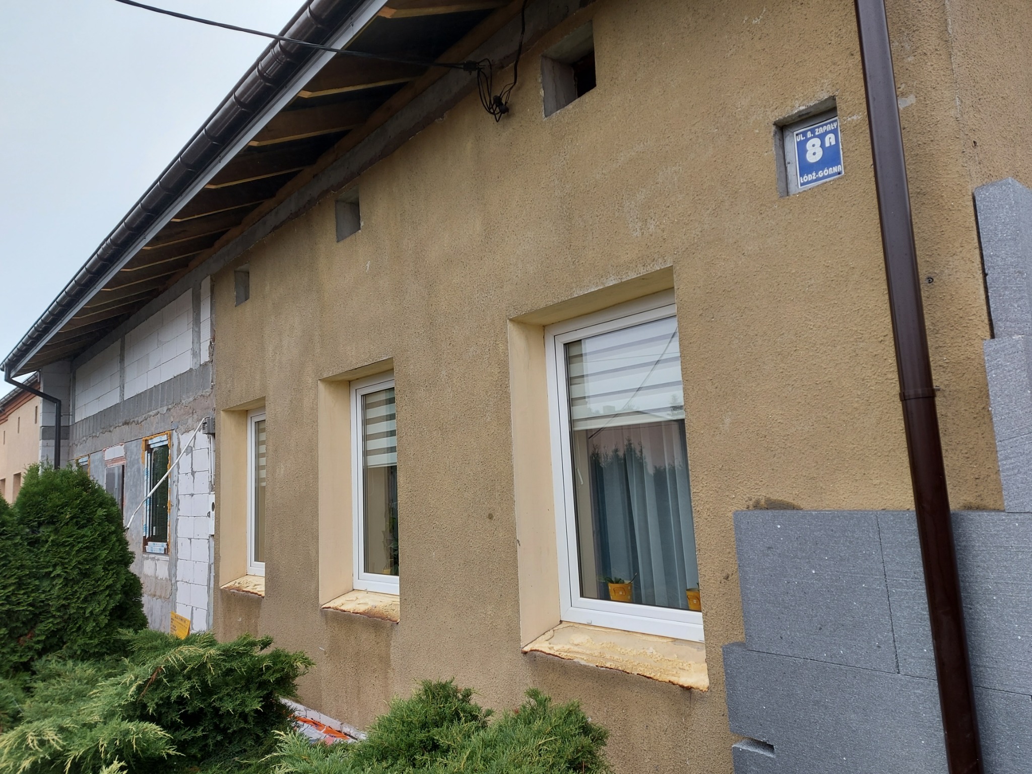 Fragment ściany budynku w trakcie prac: część otynkowana na beżowo z trzema oknami, część w trakcie docieplania szarymi płytami styropianowymi, widoczny brązowy system rynnowy i roślinność
