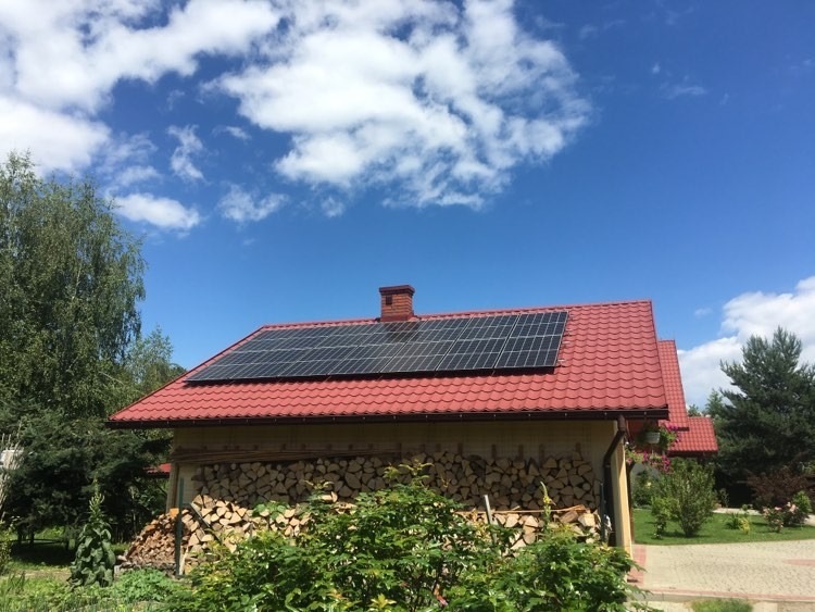 Panele słoneczne na czerwonym dachu domu jednorodzinnego, stos drewna pod ścianą, błękitne niebo z chmurami w tle.