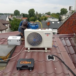 Yurii Volotkovskyi - Instalator w kraciastej koszuli montuje zewnętrzną jednostkę klimatyzacji Kaisai na brązowym dachu domu jednorodzinnego; widoczna pompa próżniowa, skrzynka narzędziowa Neo Tools i otwarte okno...