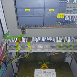 Yurii Volotkovskyi - Wnętrze szafy sterowniczej z modułami Siemens Simatic S7-1200, okablowaniem oznaczonym etykietami, listwami zaciskowymi i zasilaczem Sinamics, widoczne żółte oznaczenia na elementach.