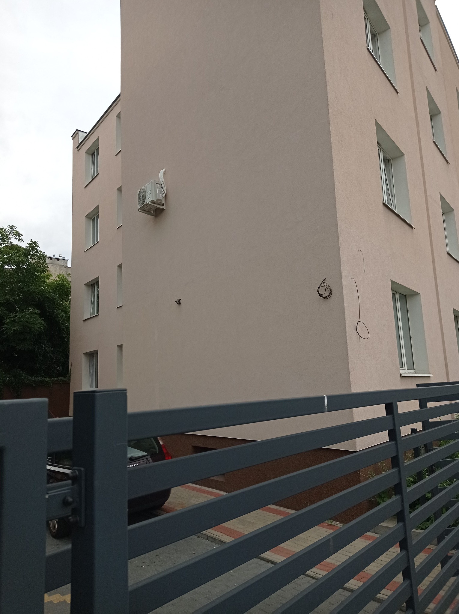 Zewnętrzna jednostka klimatyzacji zamontowana na elewacji budynku wielorodzinnego, widok zza metalowego ogrodzenia.