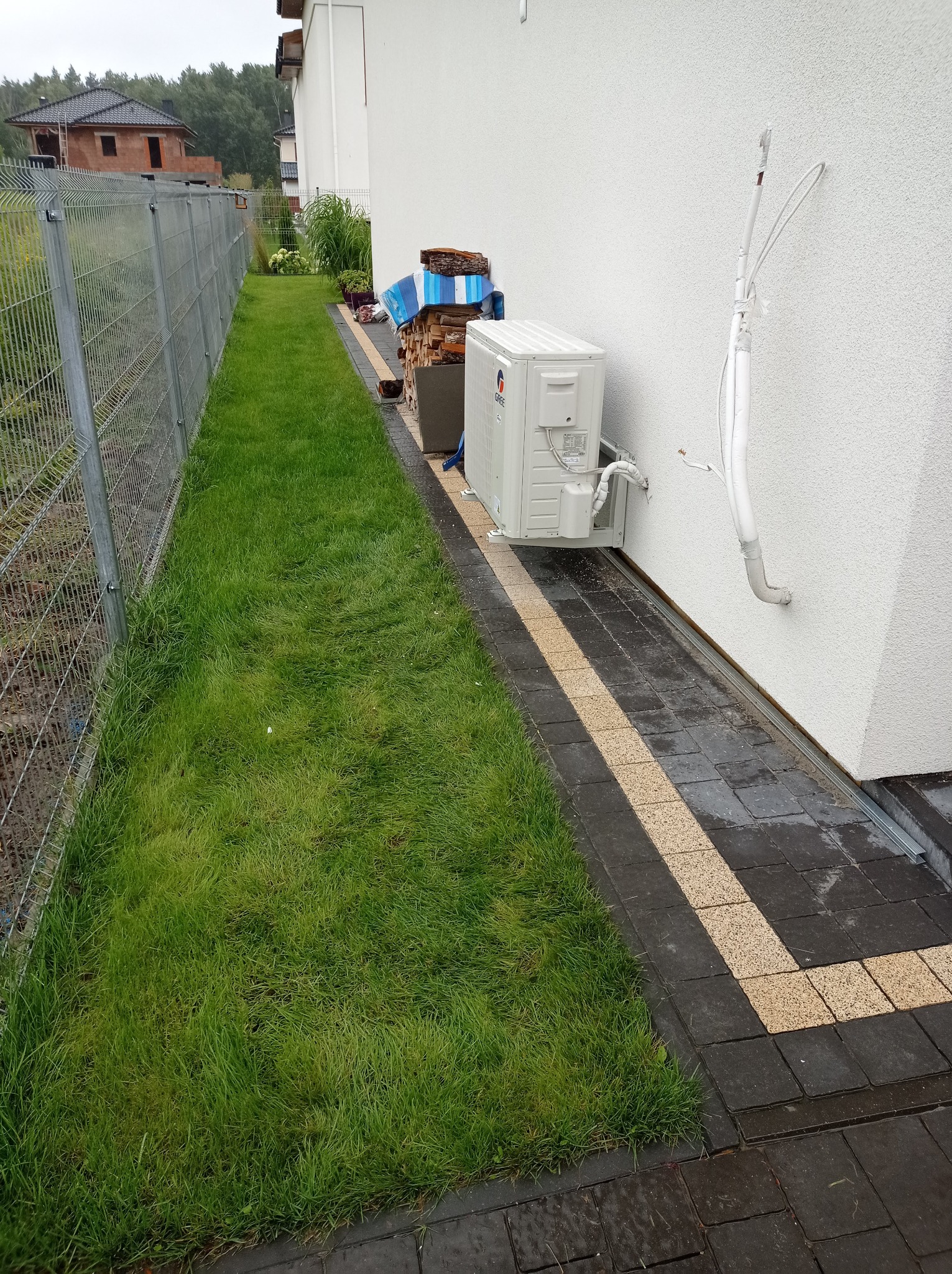Biały klimatyzator zewnętrzny zamontowany na ścianie domu, widoczne przewody instalacyjne, obok ułożone drewno kominkowe, trawnik i ogrodzenie panelowe.