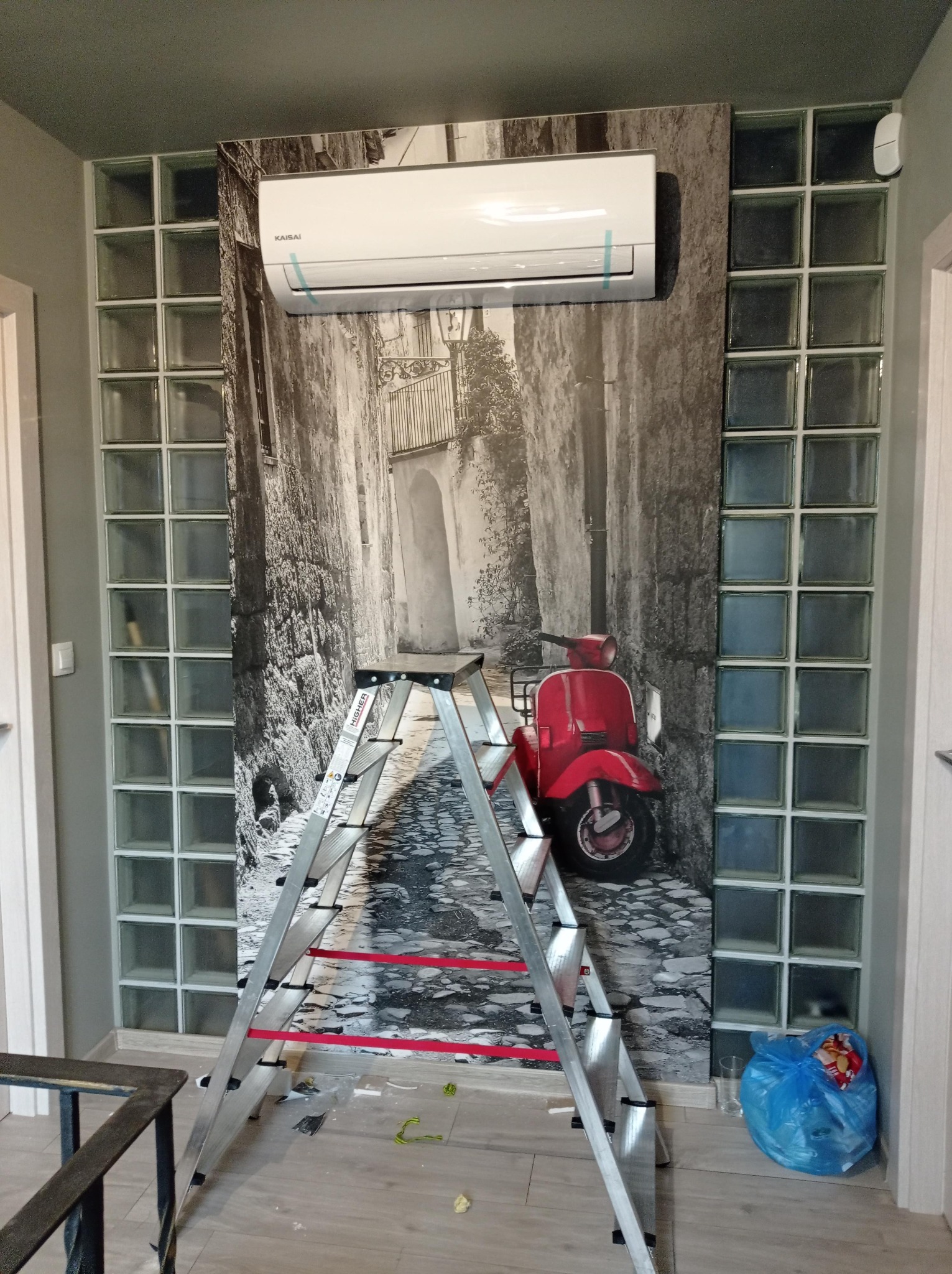 Biały klimatyzator marki Kaisai zamontowany na ścianie z fototapetą przedstawiającą uliczkę z czerwoną skuterem, widoczna aluminiowa drabina rozstawiona w pomieszczeniu z luksferami.