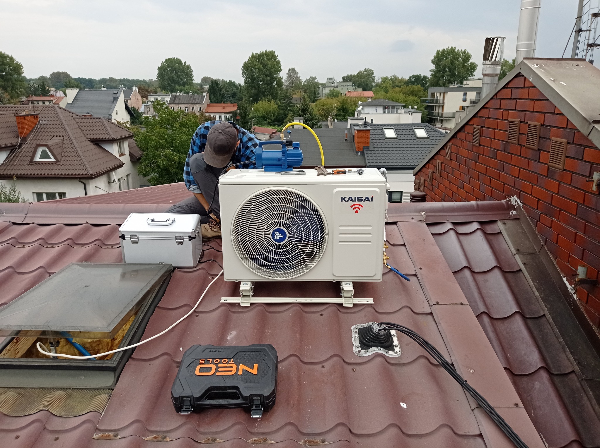 Instalator w kraciastej koszuli montuje zewnętrzną jednostkę klimatyzacji Kaisai na brązowym dachu domu jednorodzinnego; widoczna pompa próżniowa, skrzynka narzędziowa Neo Tools i otwarte okno...