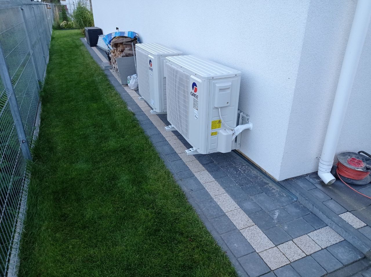 Dwa białe klimatyzatory marki Gree zamontowane na ścianie budynku, widok z zewnątrz, trawnik i kostka brukowa w otoczeniu.