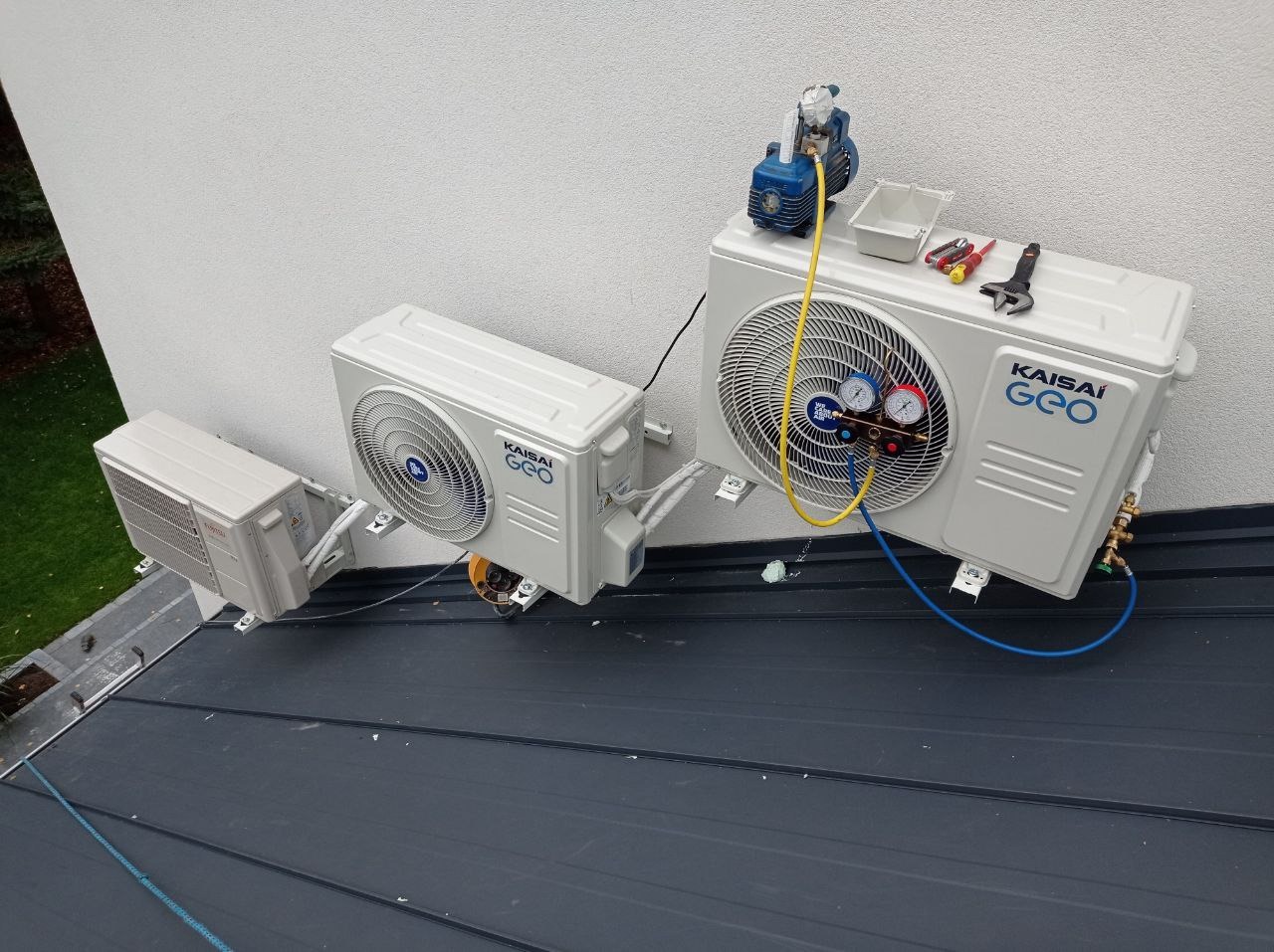 Dwa białe, zamontowane na dachu, zewnętrzne moduły klimatyzacji Kaisai Geo, podłączone do manometru i pompy próżniowej, z narzędziami obok, na tle białej ściany budynku.