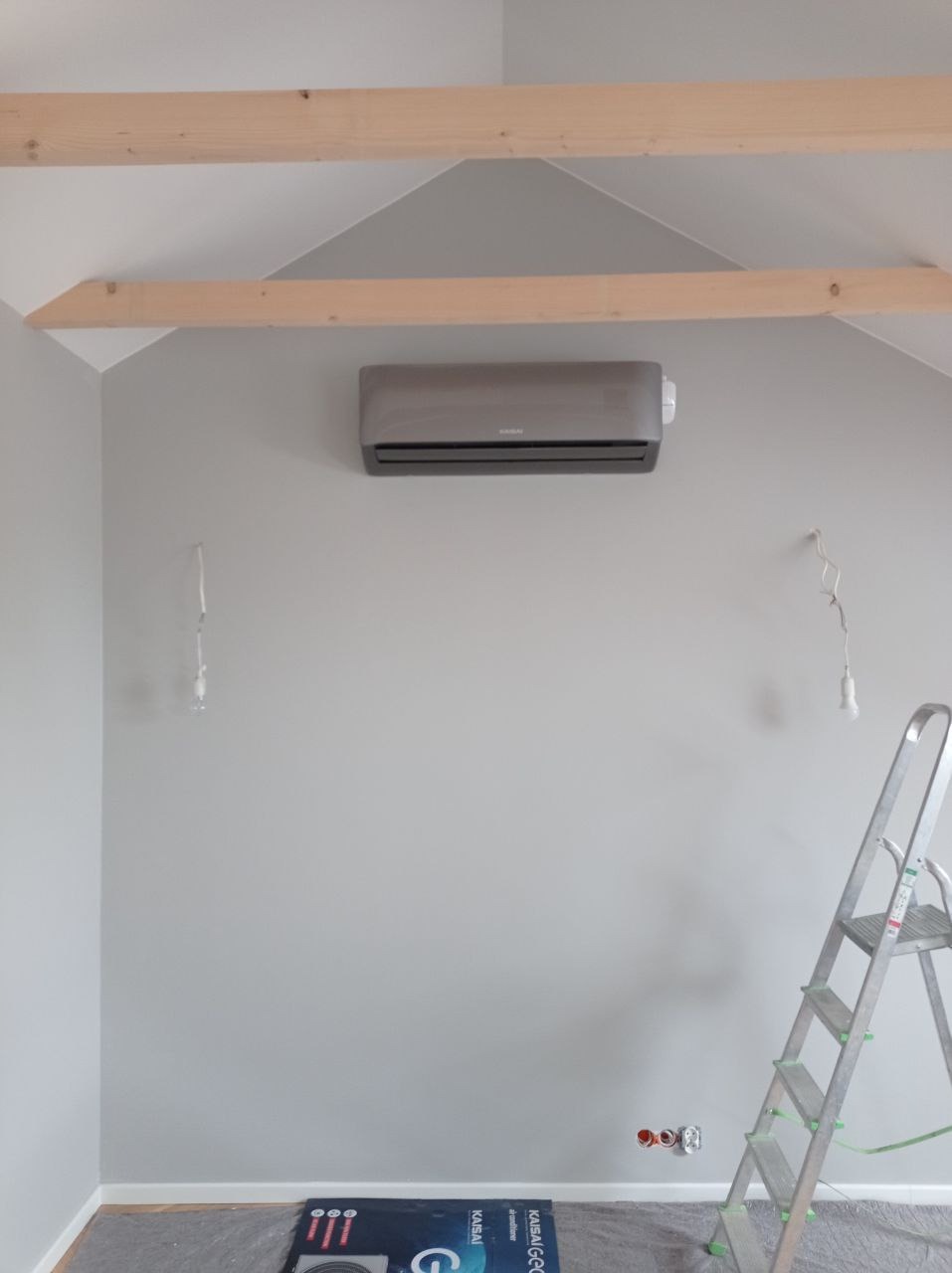 Szary klimatyzator zamontowany na szarej ścianie pod skośnym sufitem z widocznymi drewnianymi belkami, widoczne wystające kable, drabina oparta o ścianę i kartonowe opakowanie klimatyzatora...