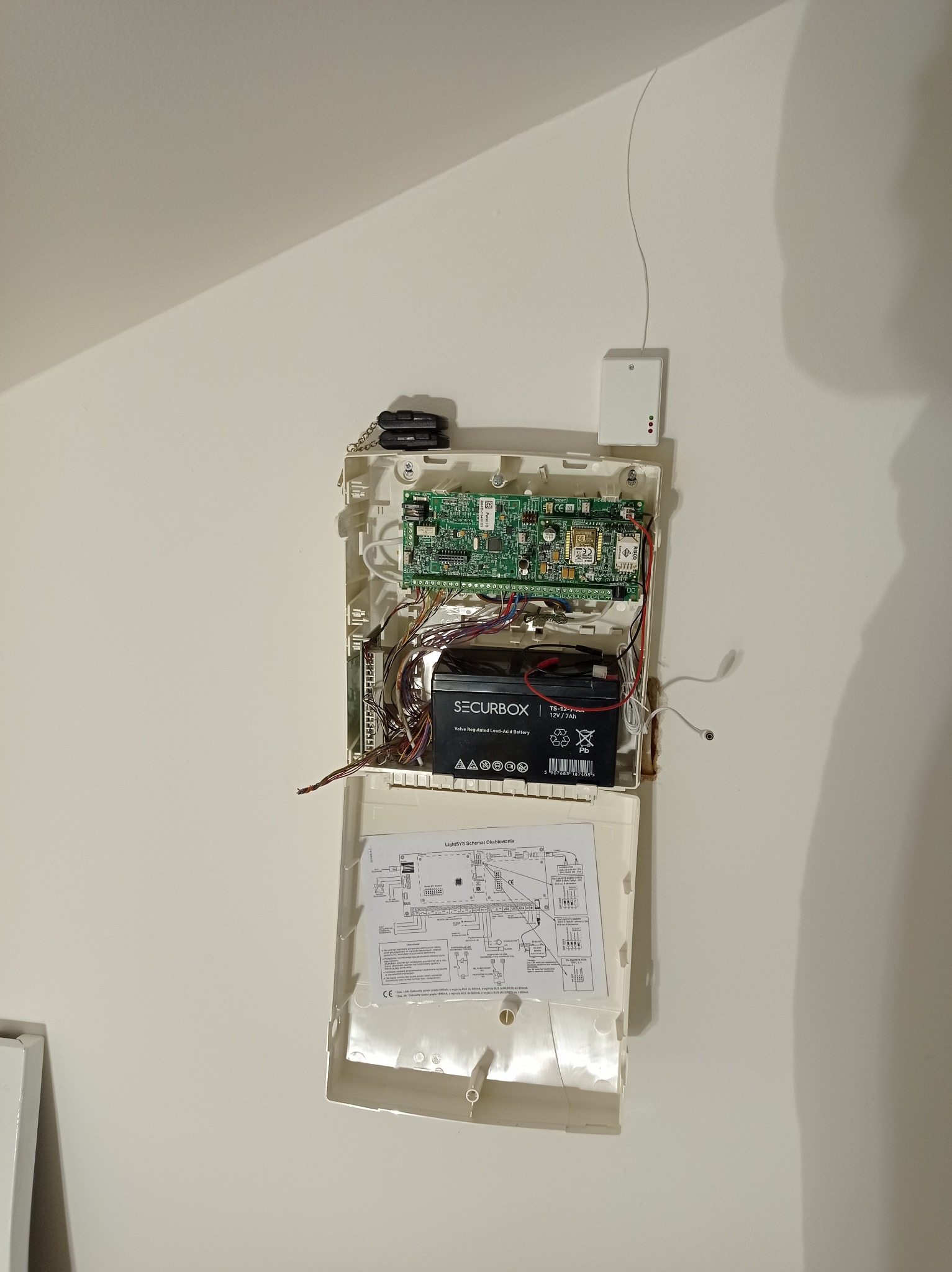 Otwarta centrala alarmowa z widoczną elektroniką, akumulatorem SECURBOX i schematem okablowania LightSYS, zamontowana na białej ścianie. Widoczny także mały pilot do alarmu i czujnik.
