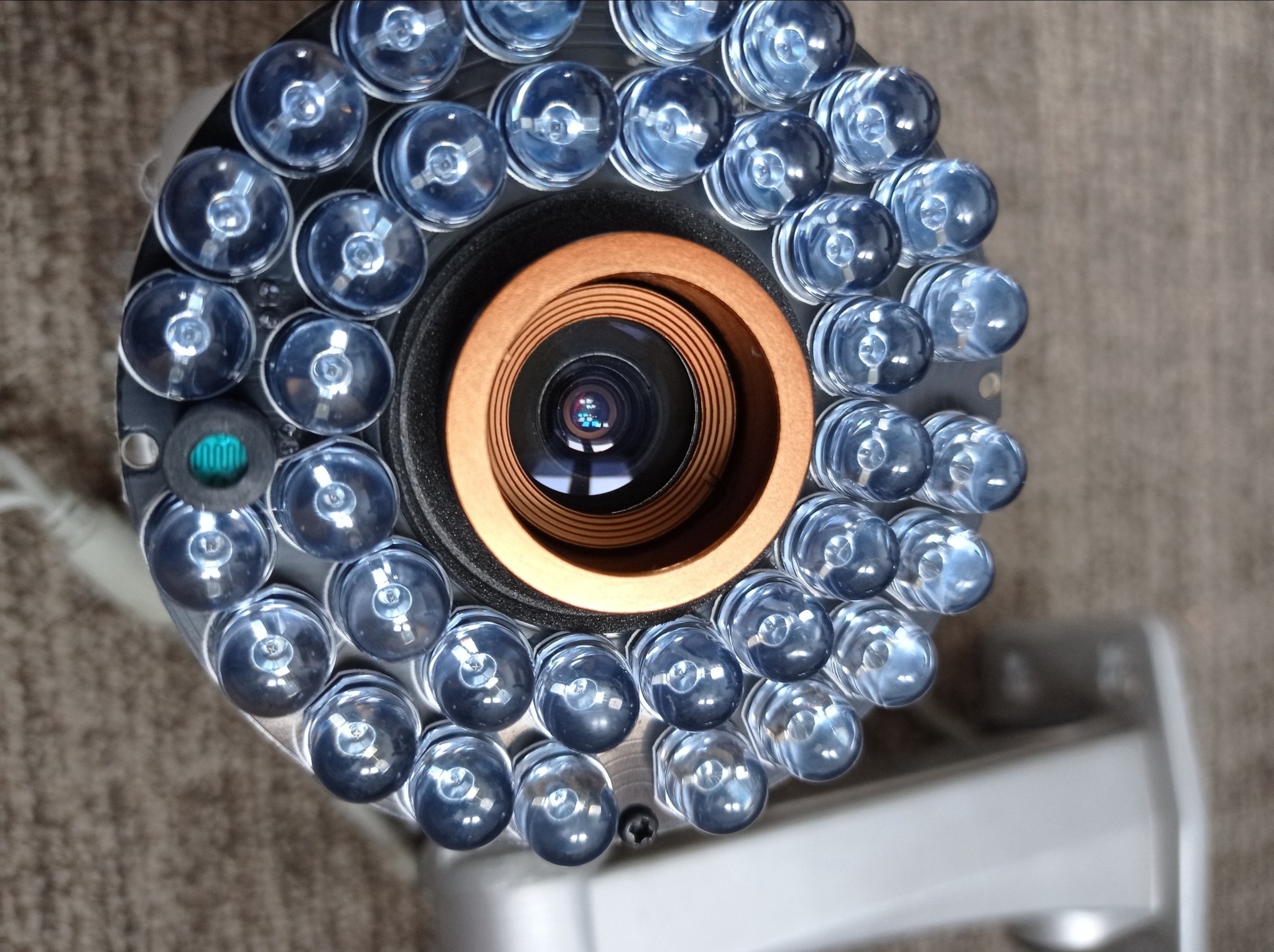 Zbliżenie na kamerę monitoringu z diodami LED, widoczny obiektyw z odbiciem światła, szara obudowa i uchwyt montażowy na rozmytym tle.