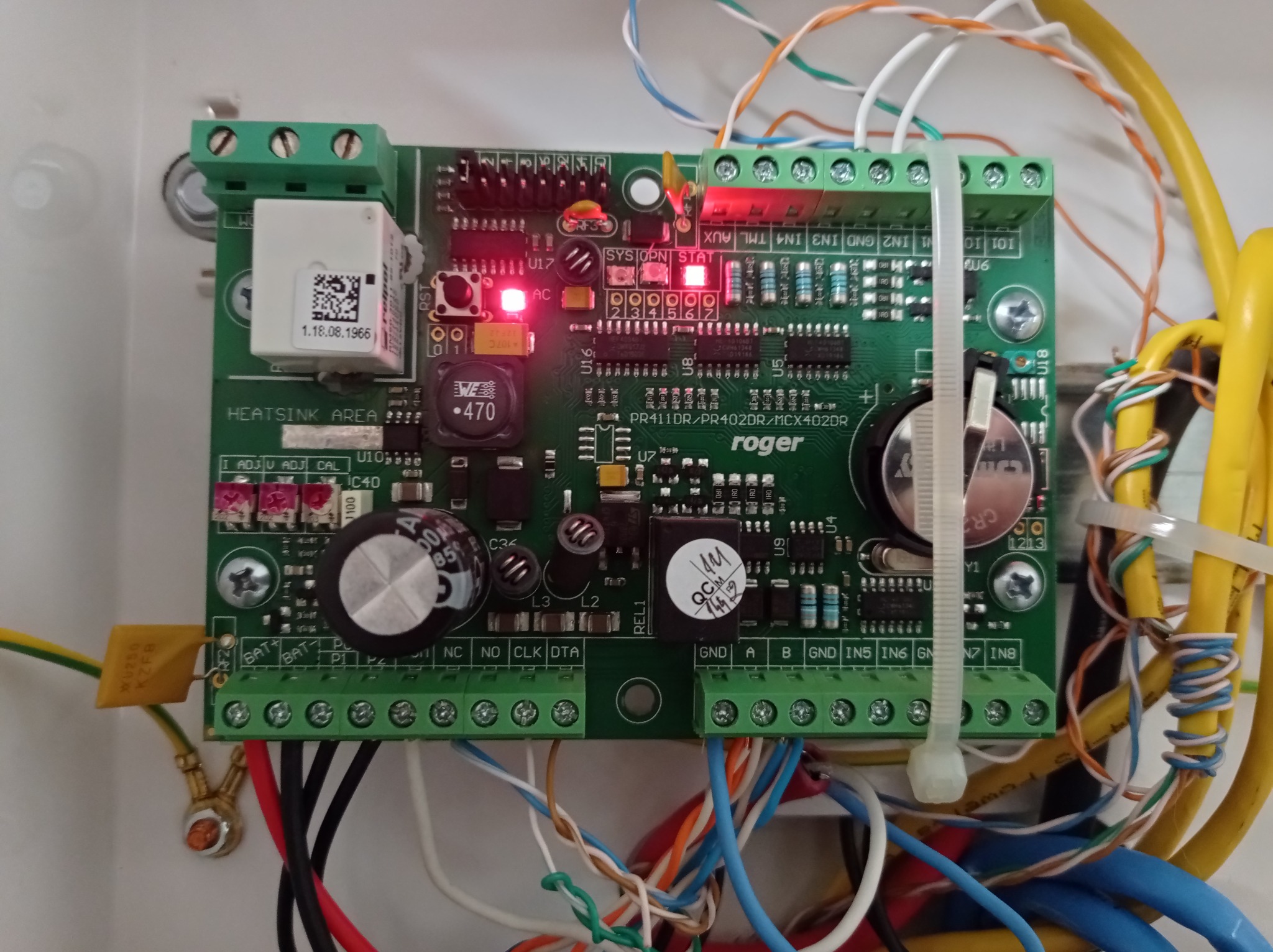 Płyta centrali alarmowej firmy Roger w białej obudowie, widoczne liczne przewody, diody LED, złącza śrubowe i komponenty elektroniczne, w tym bateria CR2032.
