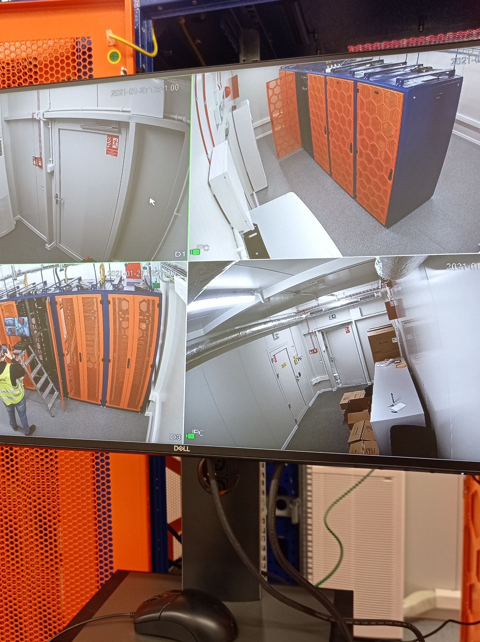 Ekran monitora z podglądem z kamer przemysłowych w serwerowni, widoczne szafy rack z pomarańczowymi panelami i pracownik na drabinie.