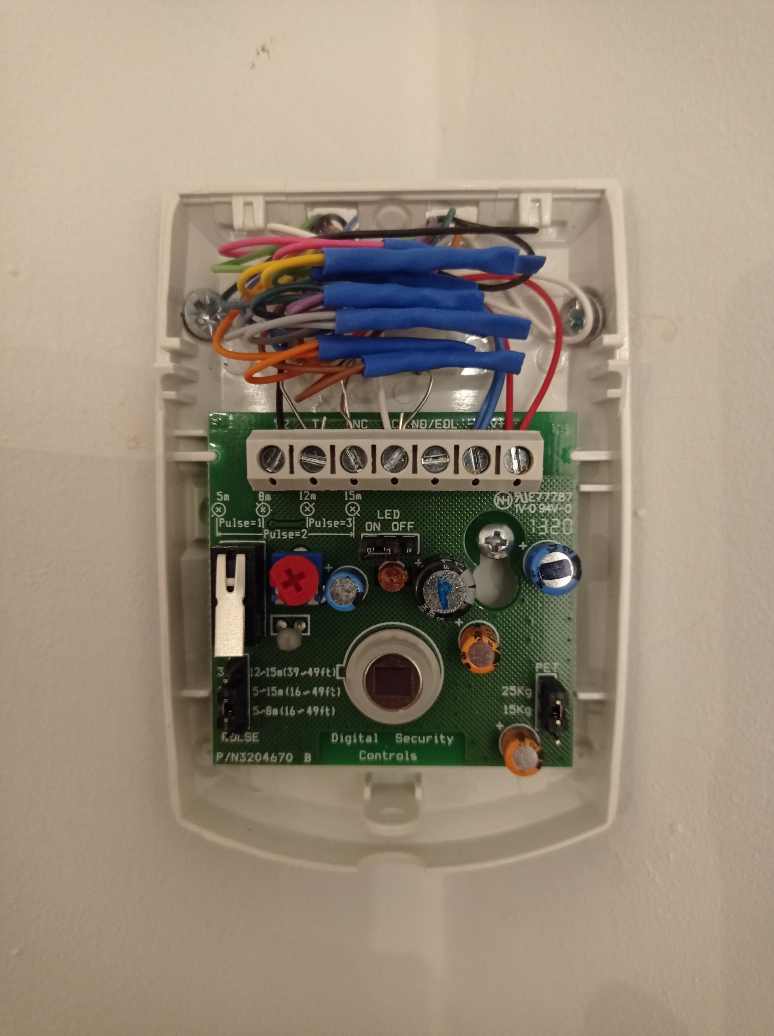 Otwarta obudowa czujnika alarmowego z widoczną płytką drukowaną, przewodami zabezpieczonymi niebieskimi złączkami i elementami elektronicznymi.