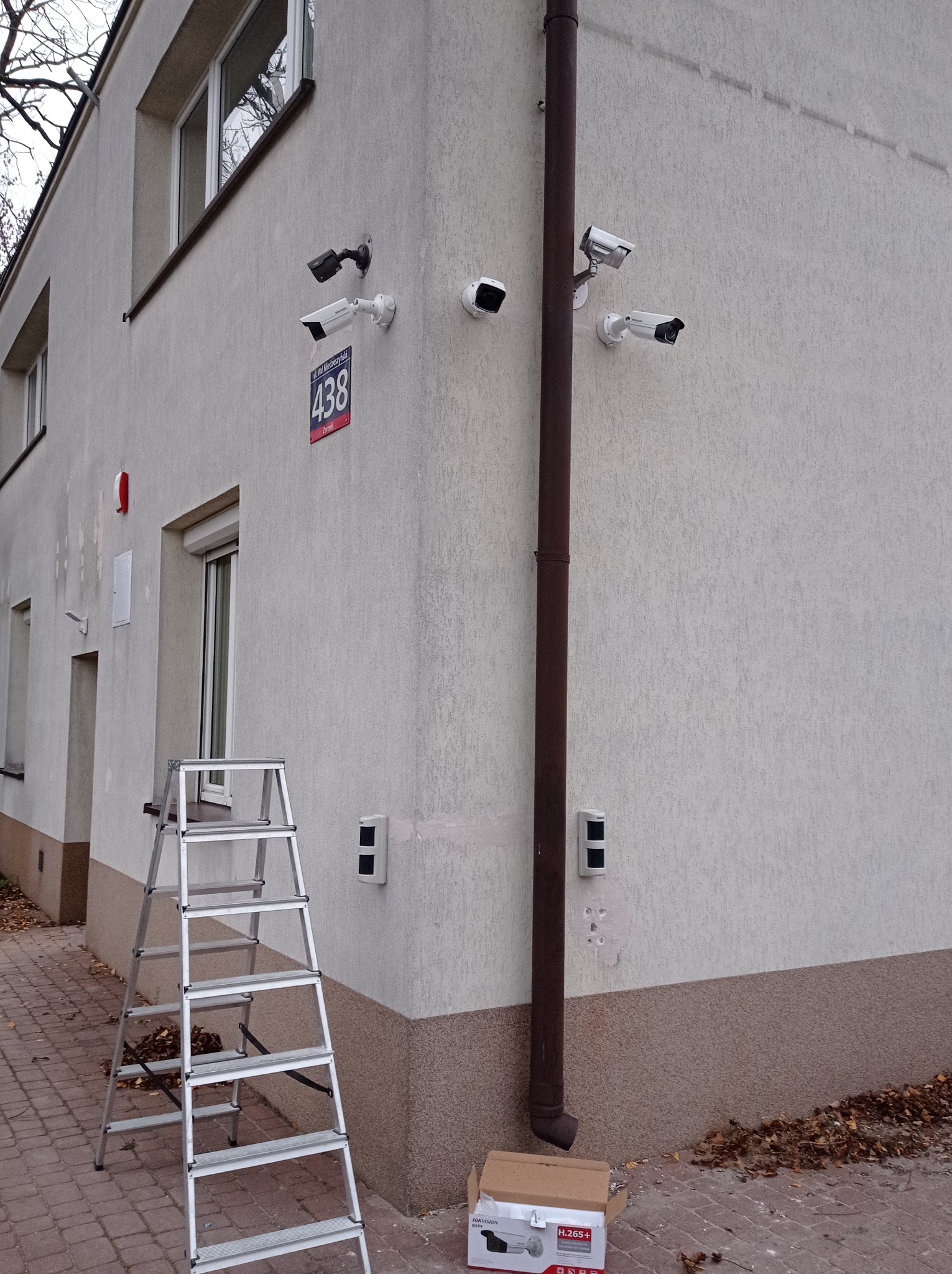 Zewnętrzny montaż systemu monitoringu: cztery kamery na elewacji budynku z numerem 438, drabina przystawiona do ściany, otwarte pudełko po kamerze na chodniku.