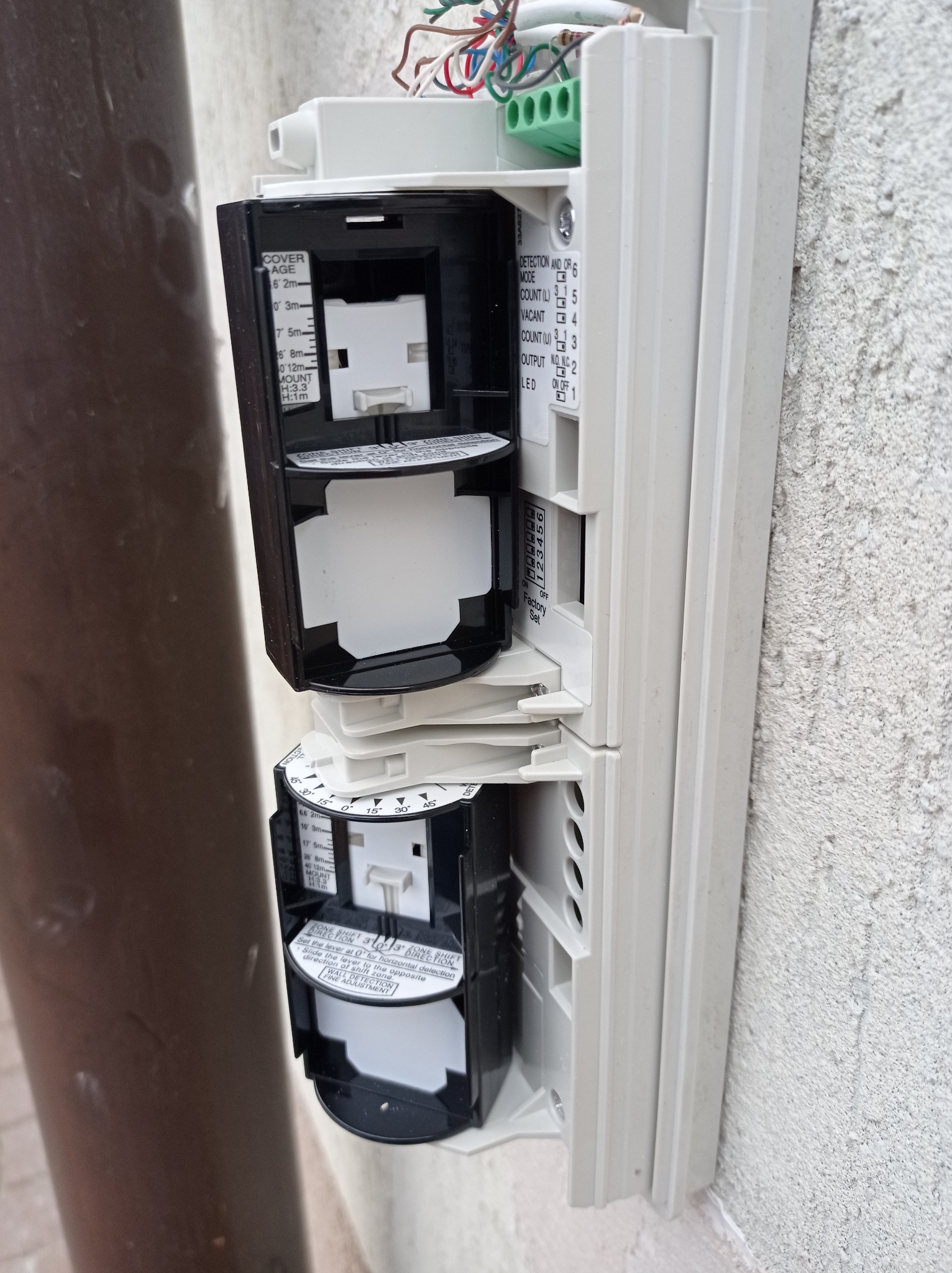 Dwa czujniki ruchu w obudowie, zamontowane na szarej, chropowatej ścianie, z widocznymi ustawieniami zasięgu i kierunku detekcji oraz przewodami elektrycznymi.