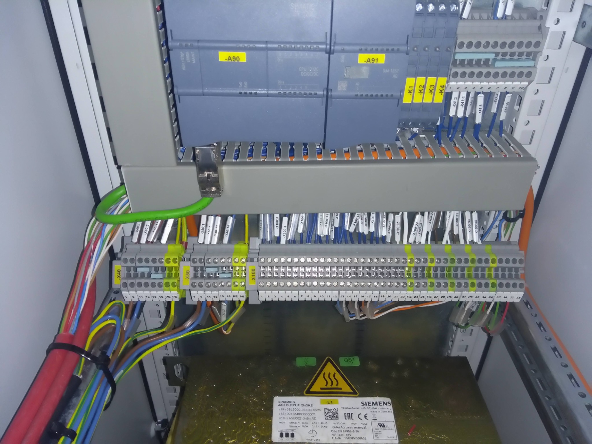 Wnętrze szafy sterowniczej z modułami Siemens Simatic S7-1200, okablowaniem oznaczonym etykietami, listwami zaciskowymi i zasilaczem Sinamics, widoczne żółte oznaczenia na elementach.