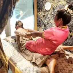 Masażystka w różowym stroju wykonuje tajski masaż na kliencie leżącym na łóżku z orientalnym wzorem, na ścianie zegar i tapeta w liście.