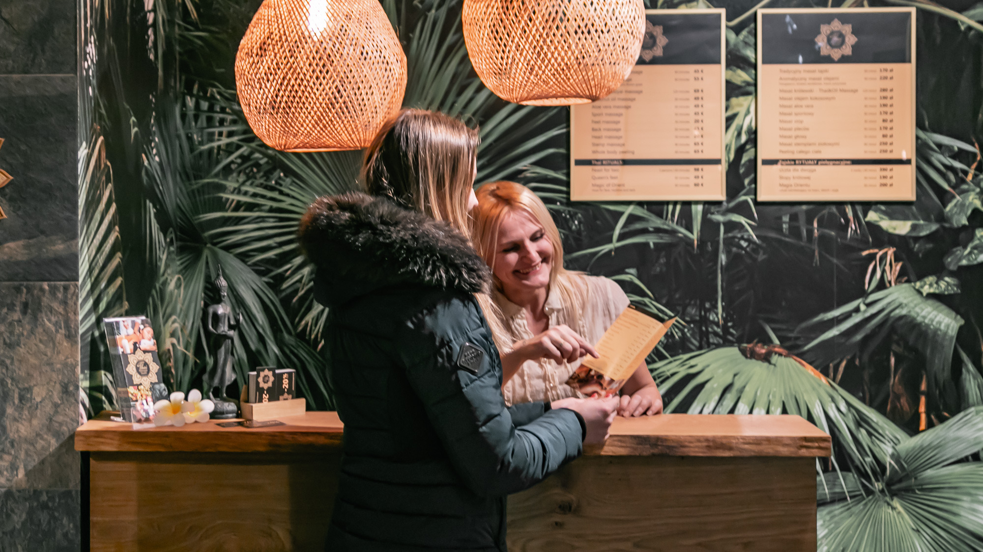 Klientka w recepcji, przegląda menu usług z uśmiechniętą pracownicą, egzotyczne dekoracje i cenniki na ścianie.