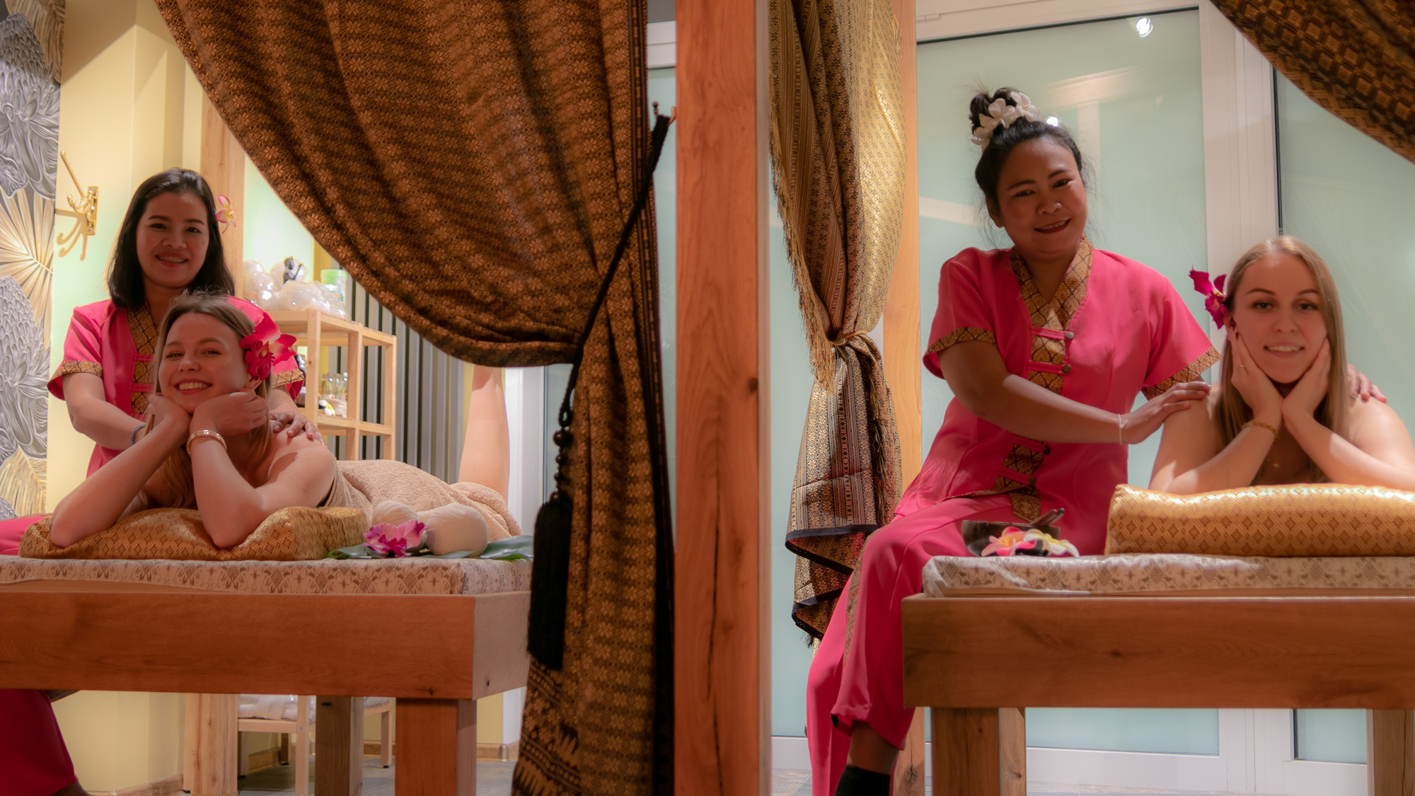 Dwie kobiety na łóżkach do masażu, jedna z nich masowana przez masażystkę w różowym uniformie, druga masażystka siedzi obok drugiej klientki, egzotyczny wystrój wnętrza z zasłonami i kwiatami.