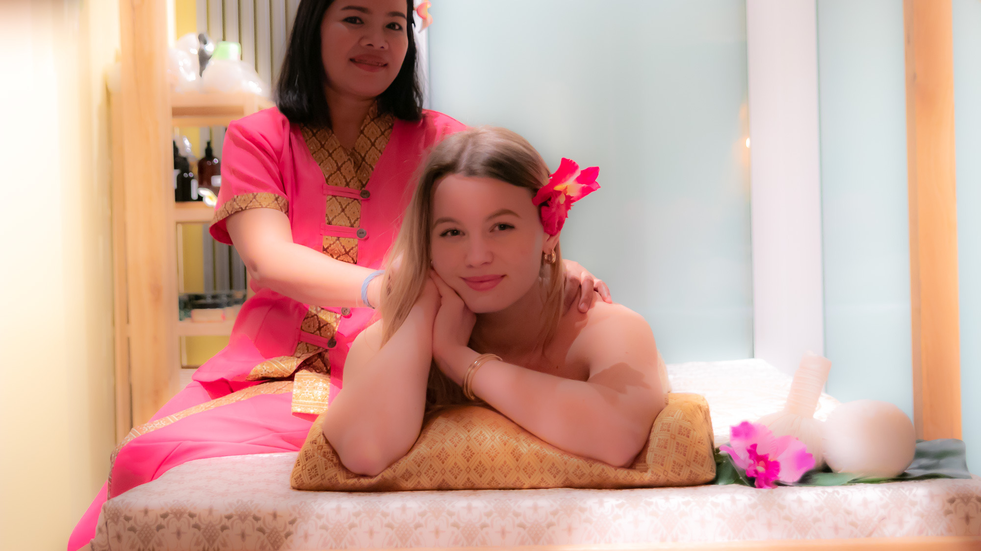 Klientka z kwiatem we włosach relaksuje się podczas masażu pleców wykonywanego przez masażystkę w tradycyjnym stroju tajskim.