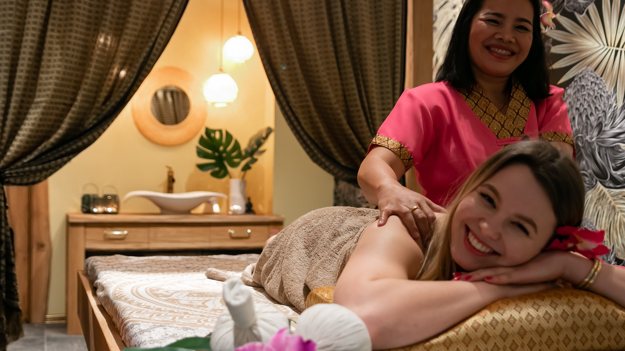 Uśmiechnięta klientka podczas masażu pleców wykonywanego przez masażystkę w różowym uniformie, w tle widoczny wystrój salonu z orientalnymi akcentami i roślinami.