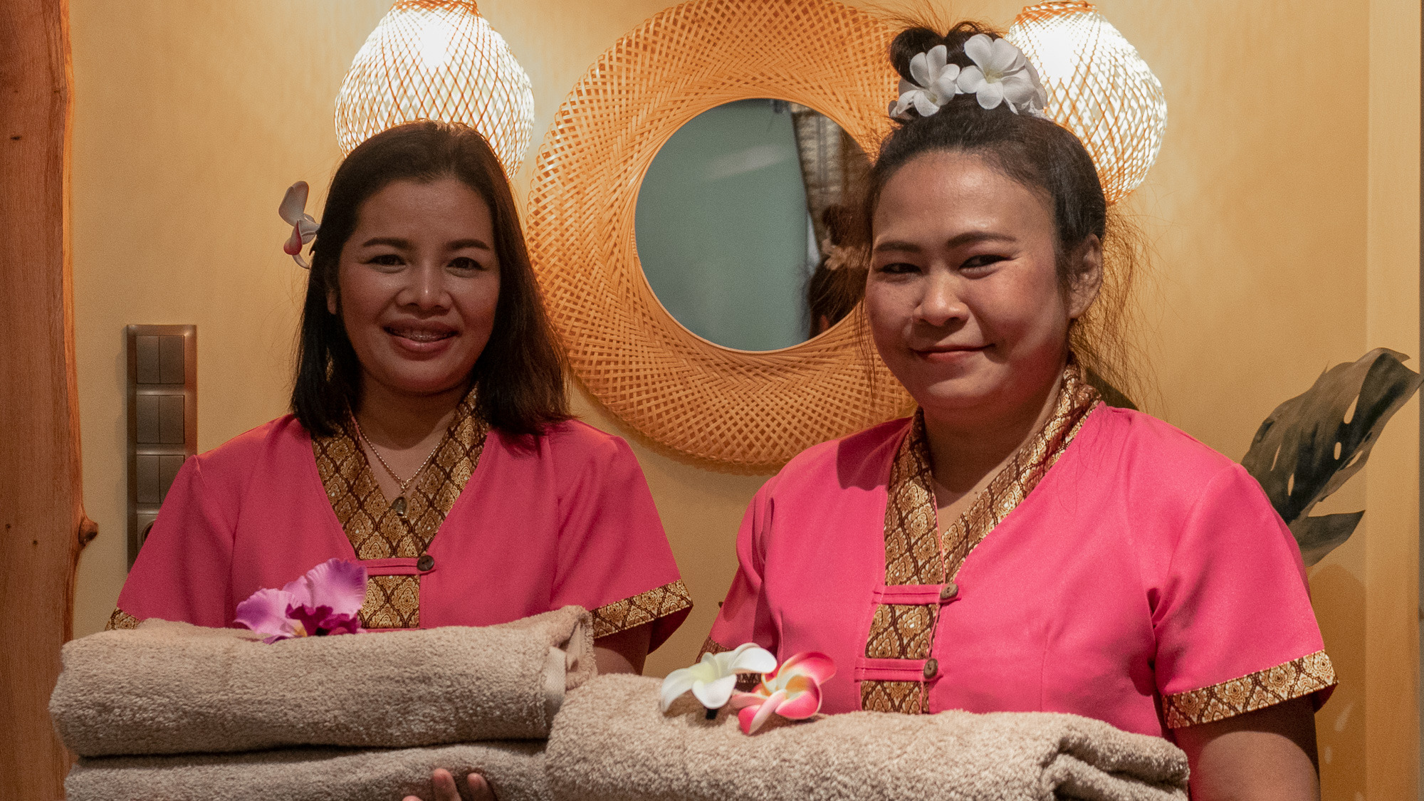 Dwie uśmiechnięte kobiety w różowych uniformach z tajskimi zdobieniami trzymają sterty ręczników w salonie z rattanowym lustrem i lampami.