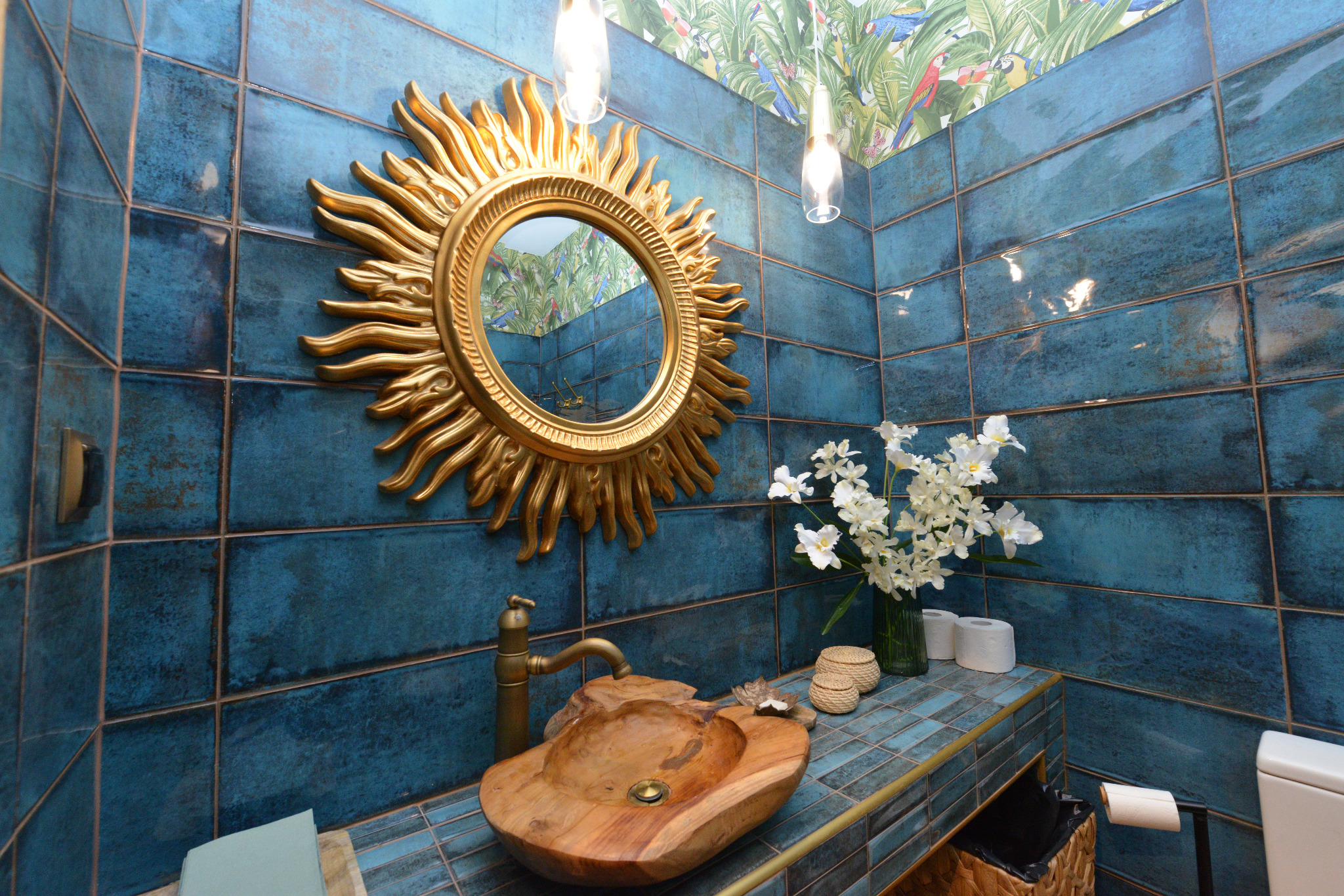 Egzotyczna łazienka z drewnianą umywalką w kształcie łodzi, złotym lustrem w kształcie słońca na tle turkusowych płytek i tapetą w tropikalne liście i papugi.