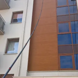 Wysoki budynek z drewnianą elewacją i oknami, czyszczony z użyciem teleskopowej lancy z szczotką i spryskiwaczem, widoczny numer budynku '4' i odbicie otoczenia w oknach parteru.