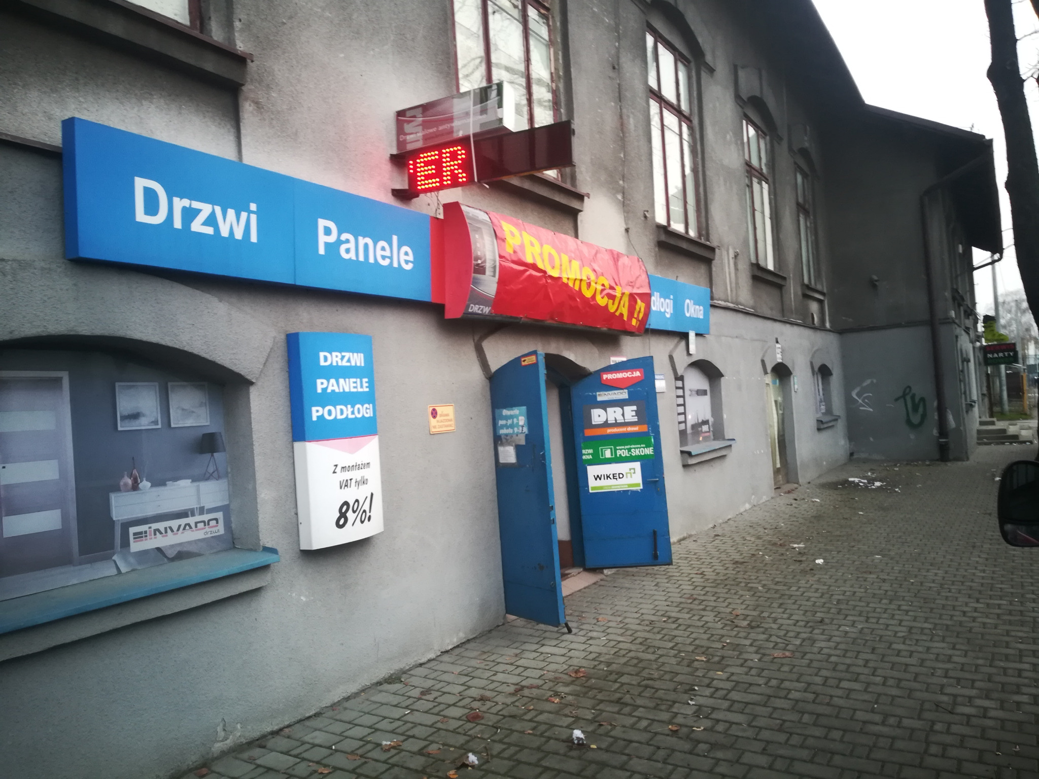 Fasada budynku z szyldami reklamującymi sprzedaż drzwi, paneli i podłóg, z otwartymi niebieskimi drzwiami wejściowymi i banerem 'PROMOCJA!' nad nimi.