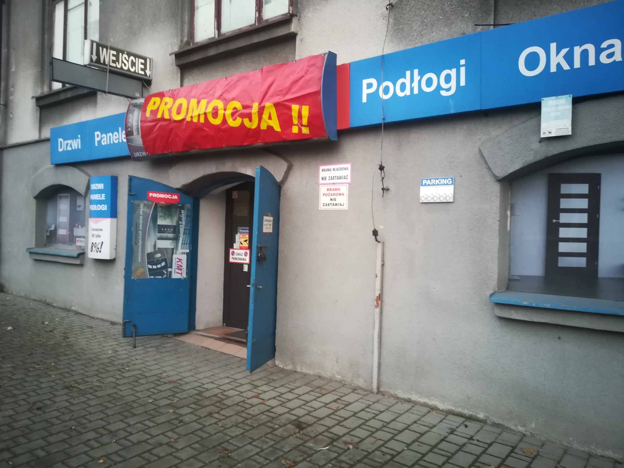 Fasada budynku z szyldami reklamowymi: Drzwi, Panele, Podłogi, Okna oraz baner 'PROMOCJA!!'. Wejście do sklepu z otwartymi, niebieskimi drzwiami.