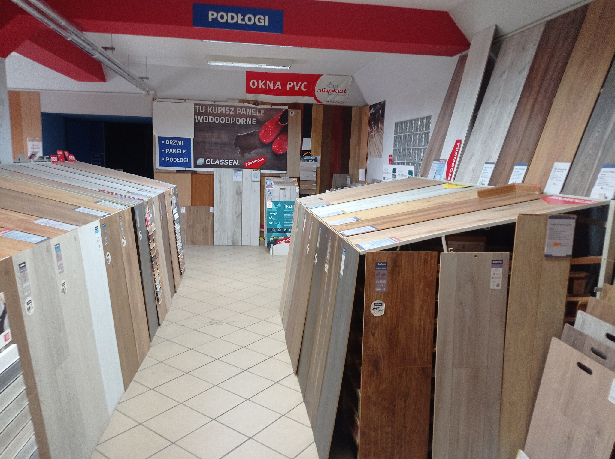 Ekspozycja różnorodnych paneli podłogowych w sklepie, widoczne także reklamy okien PVC i drzwi.