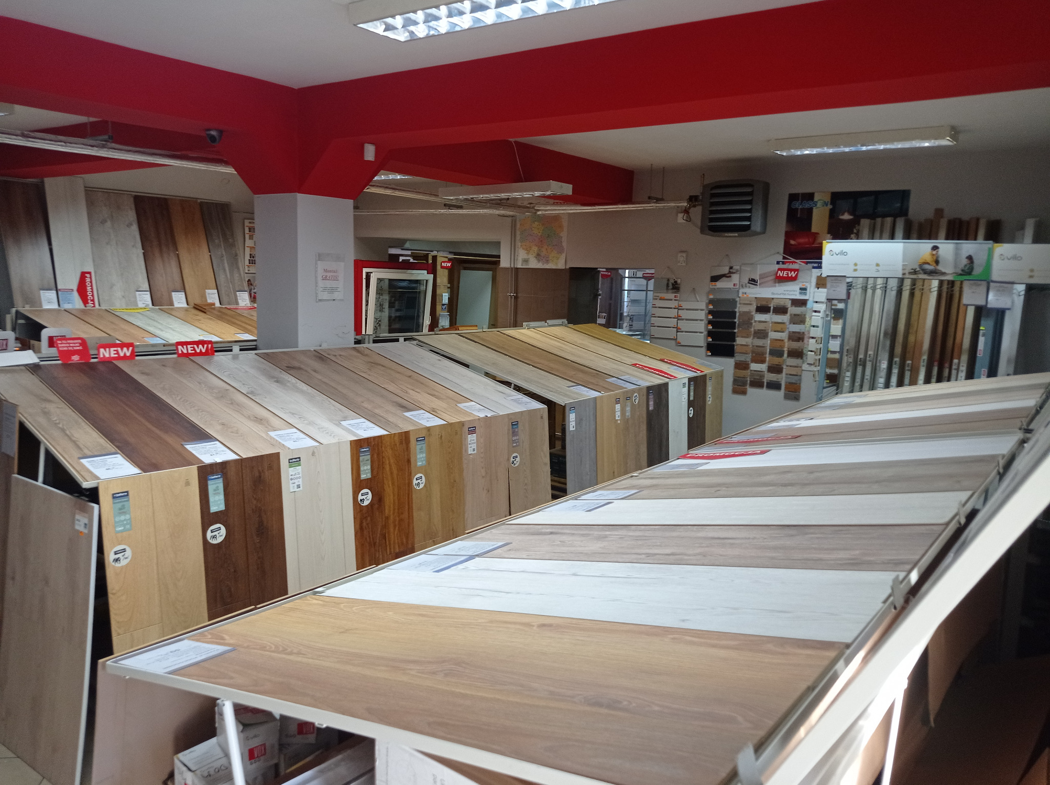 Ekspozycja różnorodnych paneli podłogowych w sklepie, prezentująca różne wzory i kolory drewna, z etykietami cenowymi i oznaczeniami 'NEW'.