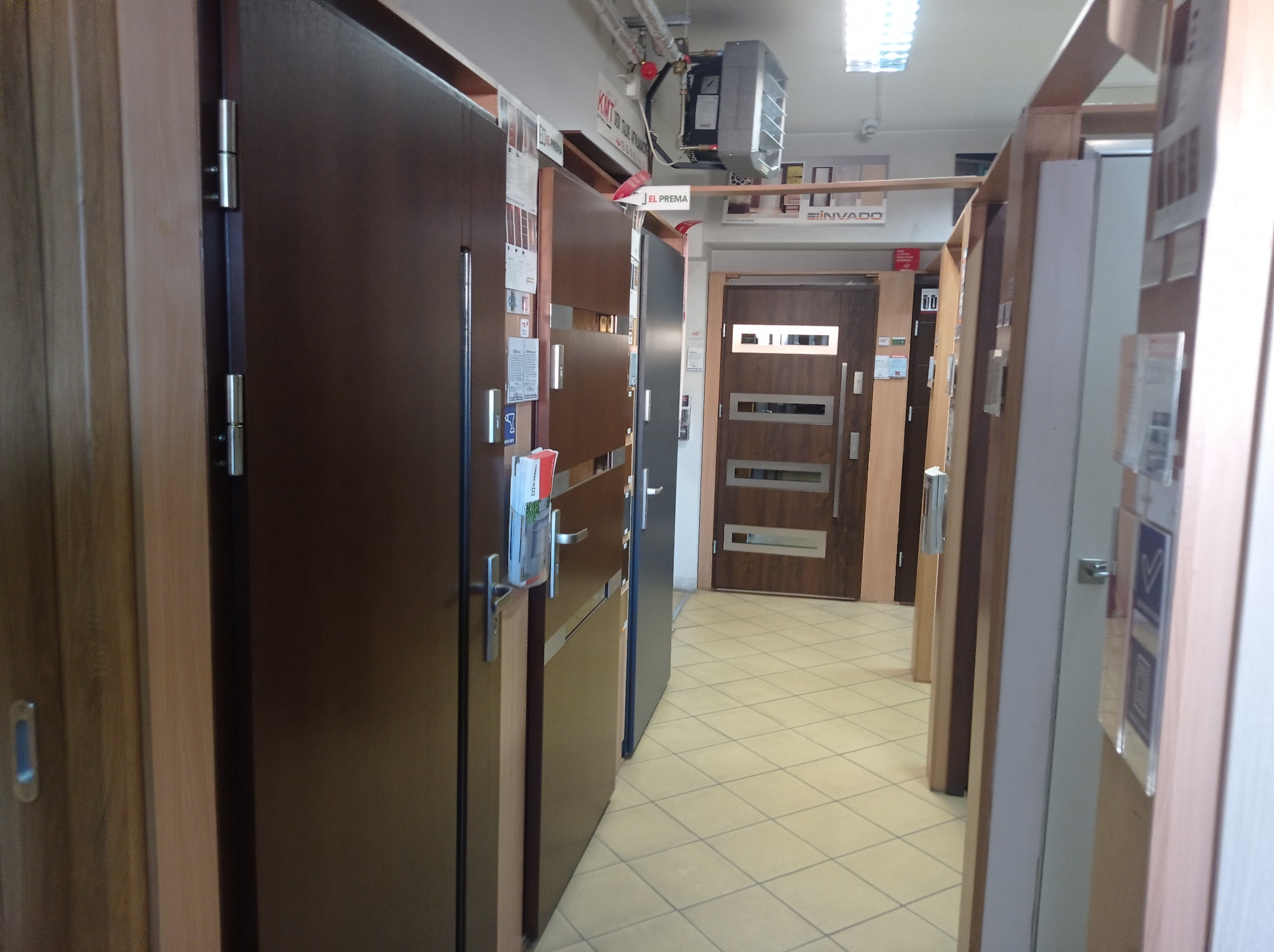 Ekspozycja różnych modeli drzwi w sklepie, widoczne drzwi drewniane i stalowe z różnymi wykończeniami i okuciami, na podłodze jasne płytki ceramiczne.