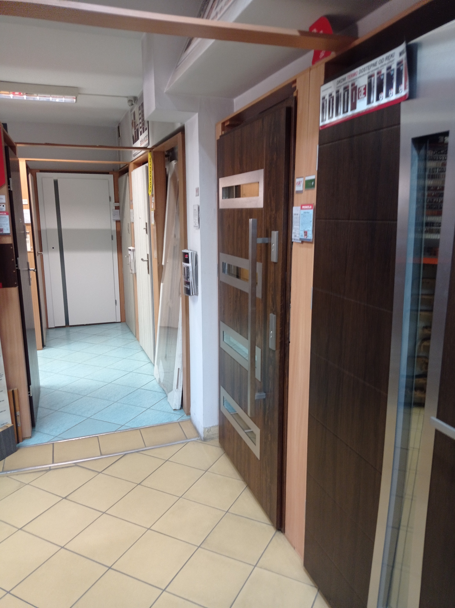 Ekspozycja różnych modeli drzwi wewnętrznych w sklepie, widoczne drzwi z przeszkleniami i metalowymi elementami dekoracyjnymi, na podłodze płytki ceramiczne.