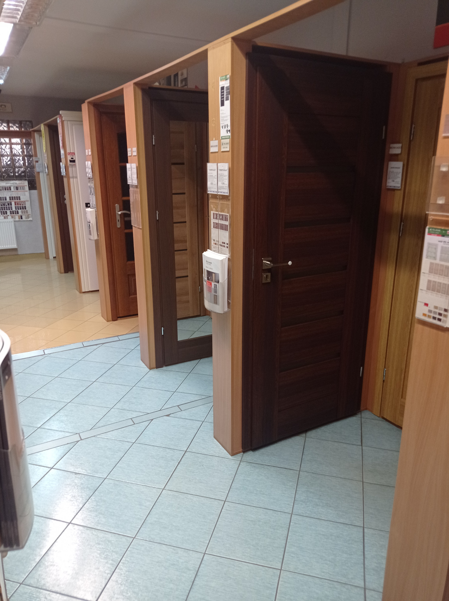 Ekspozycja różnych modeli drzwi wewnętrznych w sklepie, widoczne drzwi w odcieniach brązu i beżu z różnymi wykończeniami i klamkami, na podłodze jasne płytki.