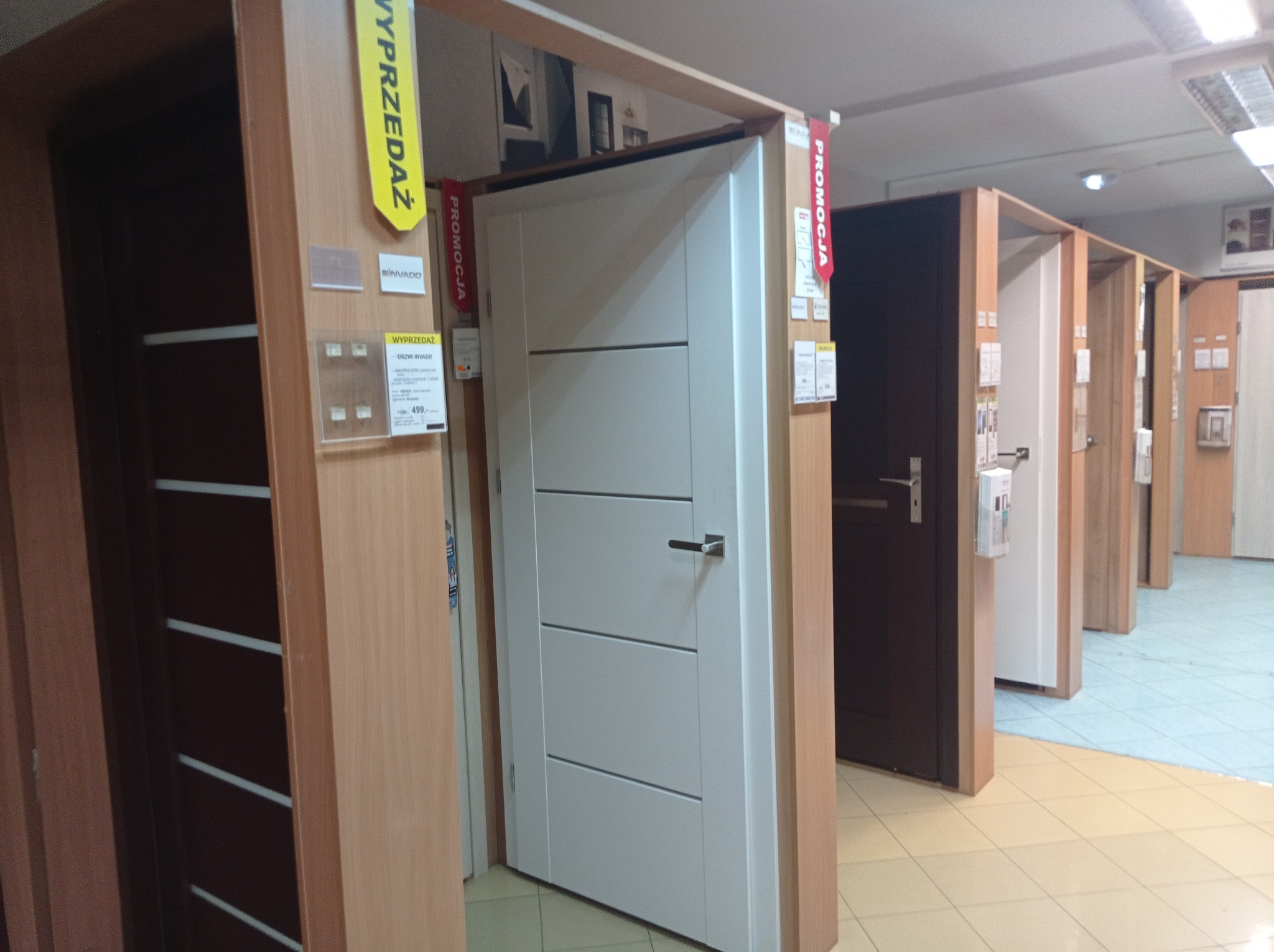 Ekspozycja wewnętrznych drzwi w sklepie: białe drzwi z poziomymi frezowaniami, obok drzwi w okleinie drewnopodobnej z żółtą etykietą 'Wyprzedaż' i czerwoną 'Promocja', widoczne inne modele w tle.