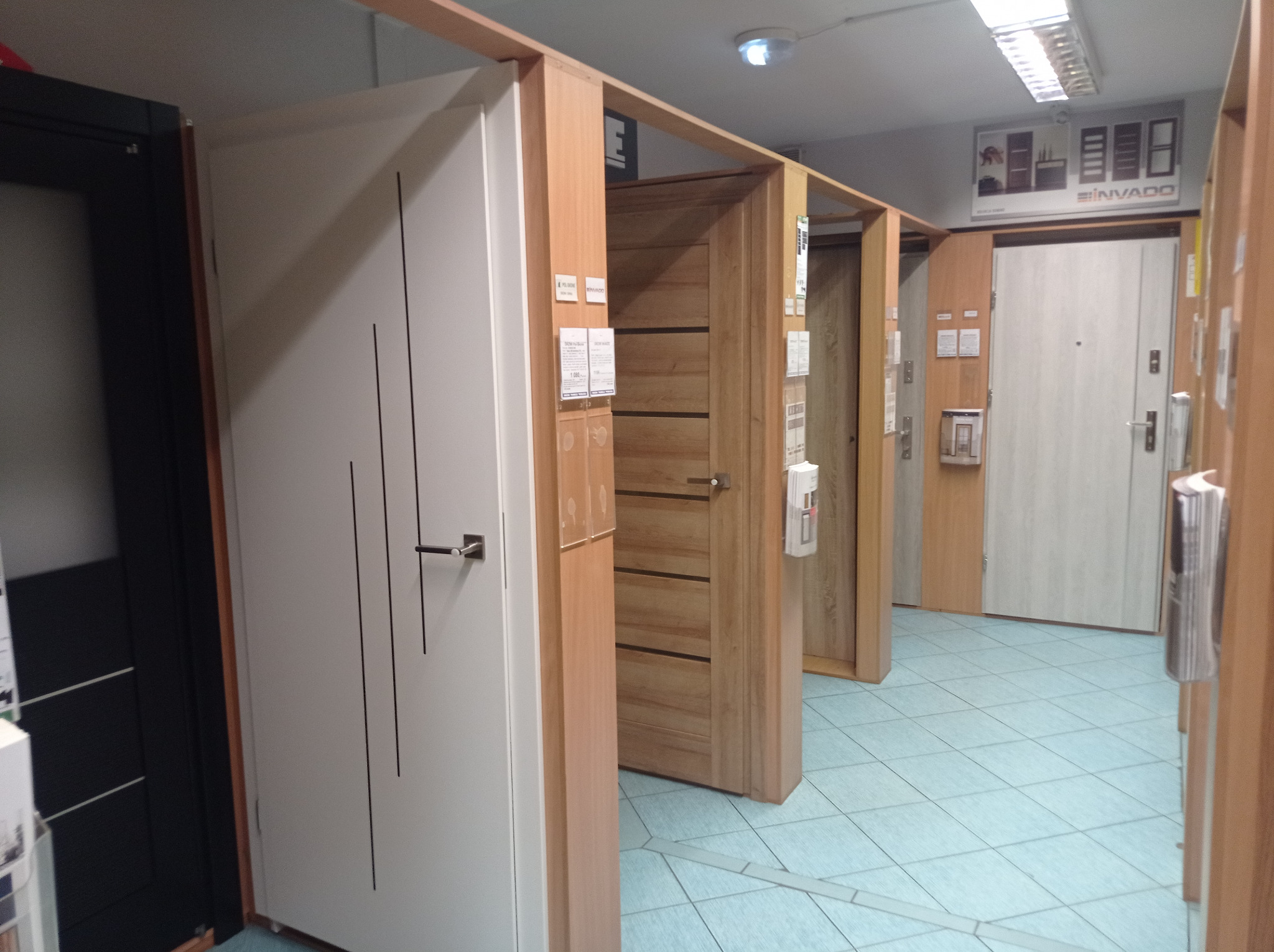 Ekspozycja różnych modeli drzwi wewnętrznych w sklepie, widoczne drzwi białe z czarnymi pionowymi paskami, drzwi drewniane z poziomymi paskami oraz drzwi białe z wizjerem