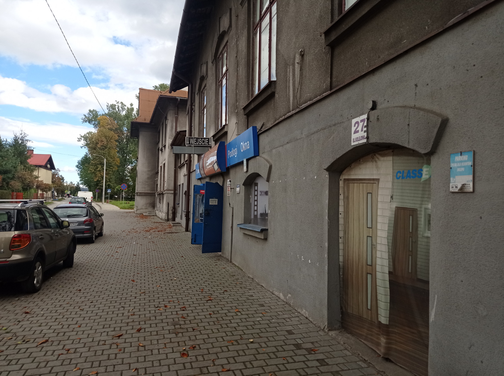 Budynek z szyldem 'Podłogi Okna' i wejściem, widok z chodnika wyłożonego kostką brukową.
