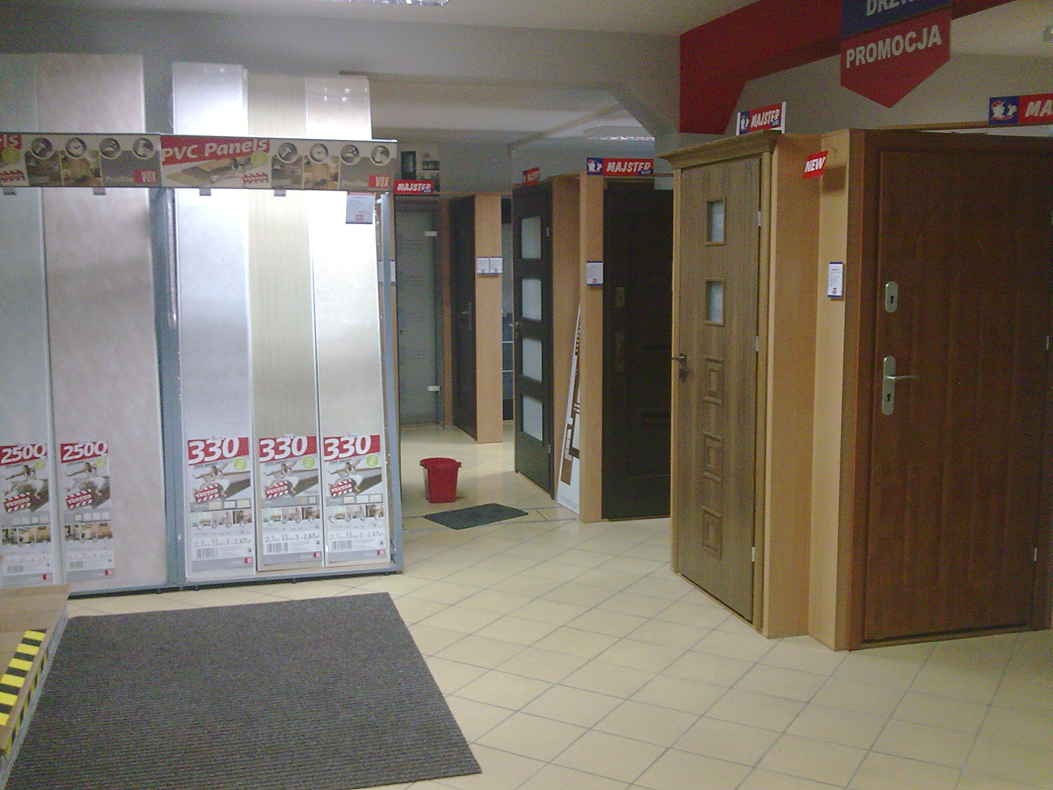 Ekspozycja różnych modeli drzwi i paneli PVC w sklepie budowlanym, widoczne oznaczenia producentów i cenówki.