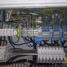 Otwarta skrzynka elektryczna z licznymi przewodami w różnych kolorach, złączkami i bezpiecznikami Siemens, widoczne elementy instalacji elektrycznej wewnątrz obudowy.