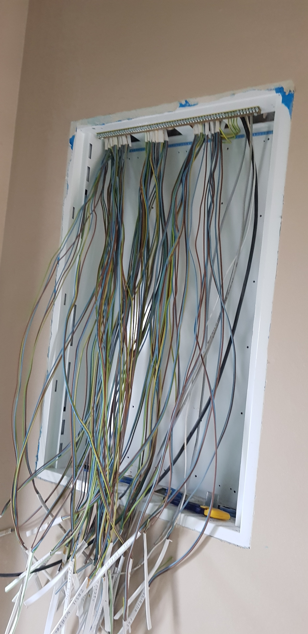 Rozdzielnica elektryczna w trakcie montażu z widocznymi przewodami w różnych kolorach, częściowo podłączonymi do listwy zaciskowej, z żółtymi obcęgami w tle.