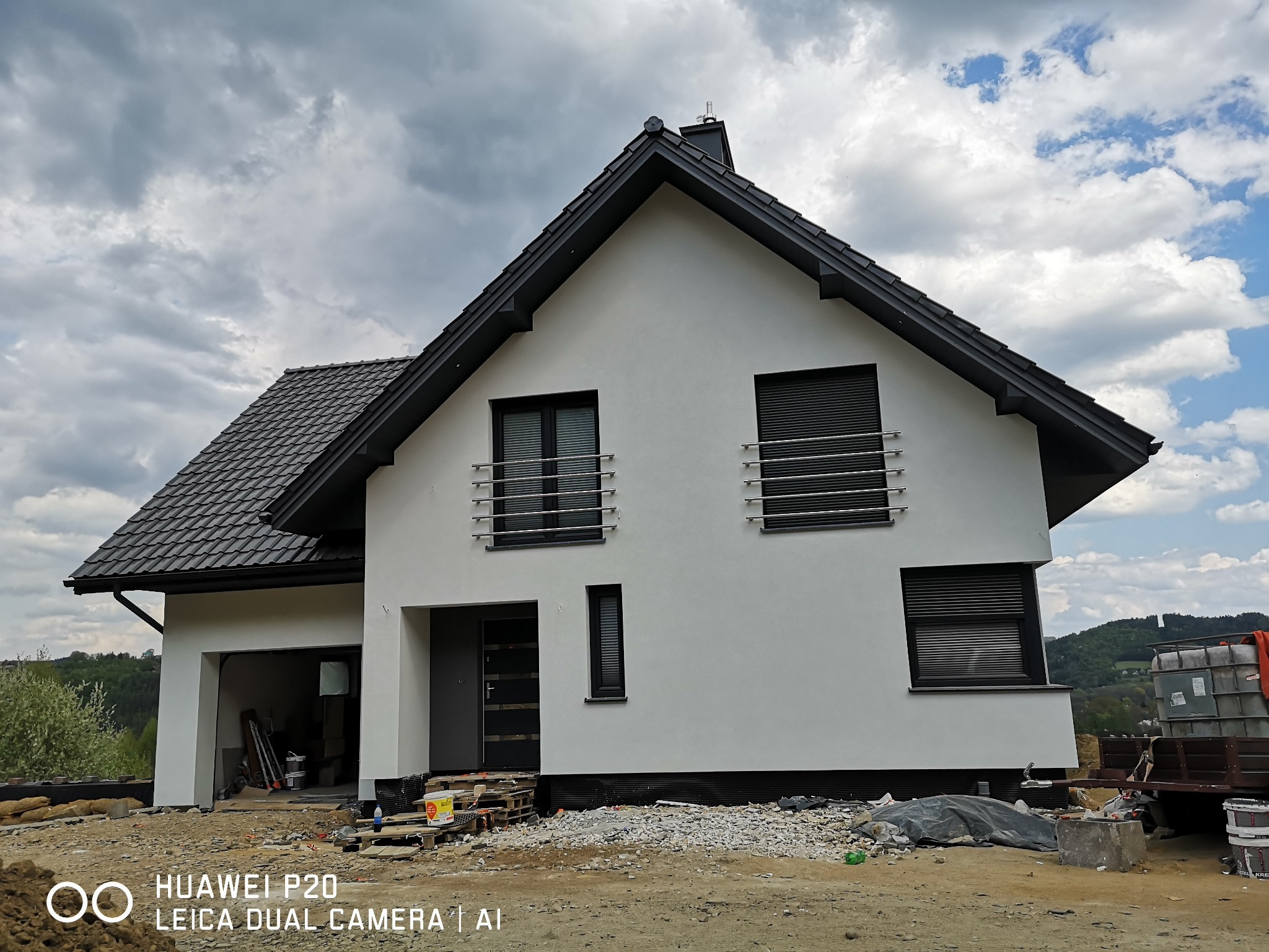 Nowoczesny dom jednorodzinny z białymi ścianami i ciemną dachówką w trakcie budowy, widoczne materiały budowlane przed budynkiem, pochmurne niebo w tle.