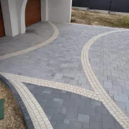 Ułożona szara kostka brukowa z jasnymi obwódkami tworzącymi dekoracyjny wzór na podjeździe przed garażem z łukowym wejściem i czarnym ogrodzeniem w tle.