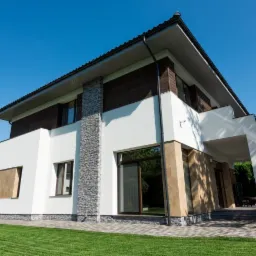 Nowoczesny dom z białą elewacją, fragmentami wykończonymi kamieniem i drewnem, widoczny z poziomu trawnika w słoneczny dzień.