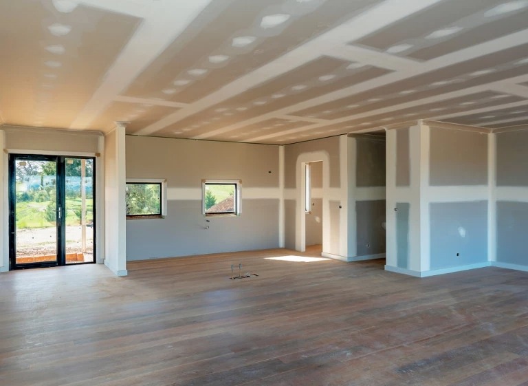 Wnętrze w stanie deweloperskim z zamontowanymi płytami gipsowo-kartonowymi na ścianach i suficie, widoczne łączenia płyt wymagające szpachlowania, drewniana podłoga i duże okno balkonowe.