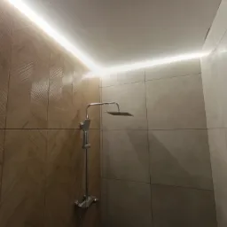 Nowoczesna kabina prysznicowa z deszczownicą i podświetleniem LED wzdłuż sufitu, wykończona płytkami w odcieniach beżu i brązu z wzorem jodełki na jednej ze ścian.