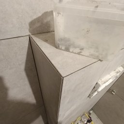 ArtMar Complex - Układanie szarych płytek w narożniku pomieszczenia w Donimierzu, widoczne plastikowe elementy do poziomowania płytek i pojemnik z klejem.