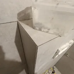 Układanie szarych płytek w narożniku pomieszczenia w Donimierzu, widoczne plastikowe elementy do poziomowania płytek i pojemnik z klejem.