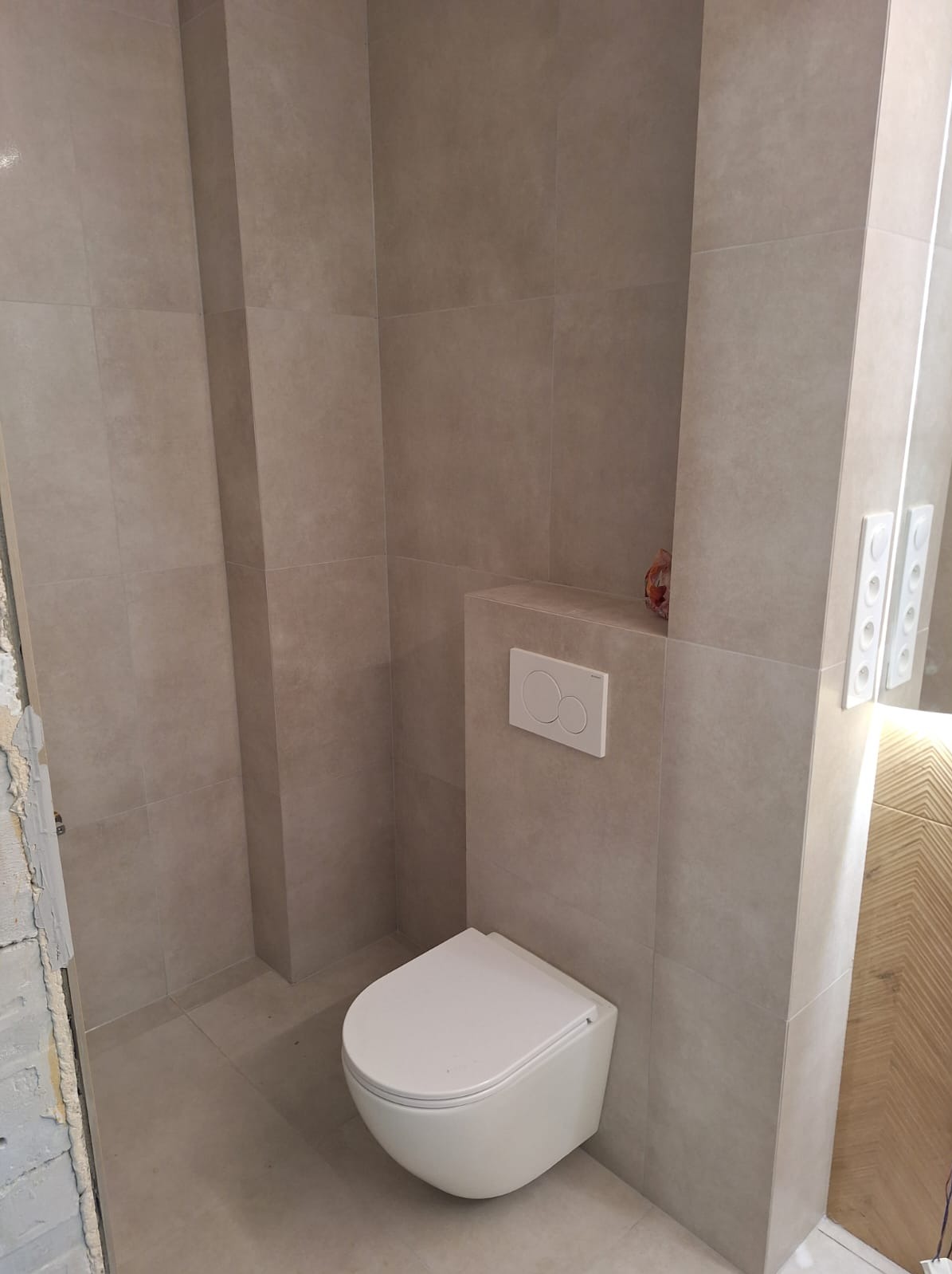 Wykończona łazienka z beżowymi płytkami, wisząca toaleta z podtynkową spłuczką, oświetlenie LED przy lustrze i częściowo wykończona ściana po lewej stronie.