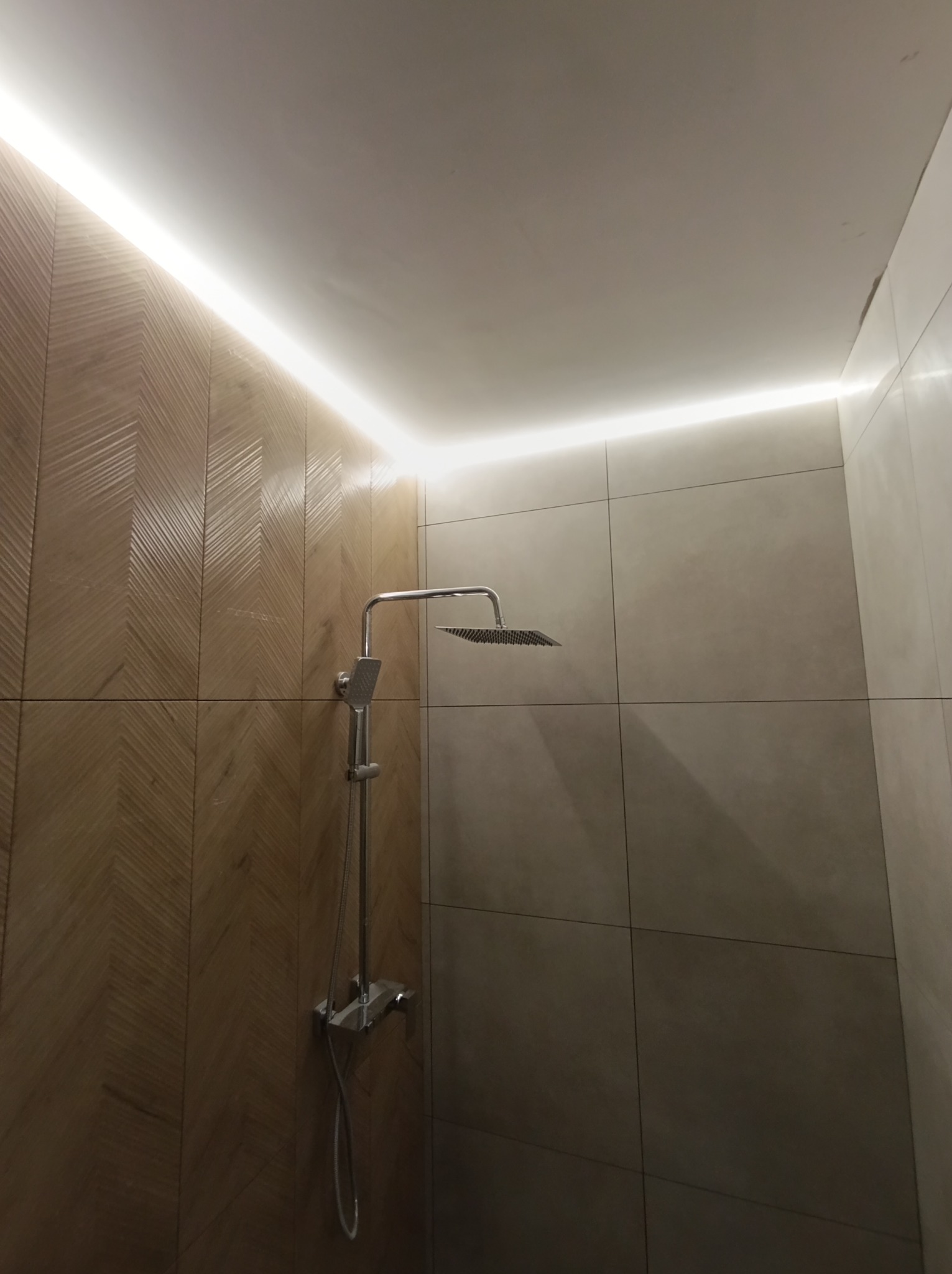 Nowoczesna kabina prysznicowa z deszczownicą i podświetleniem LED wzdłuż sufitu, wykończona płytkami w odcieniach beżu i brązu z wzorem jodełki na jednej ze ścian.