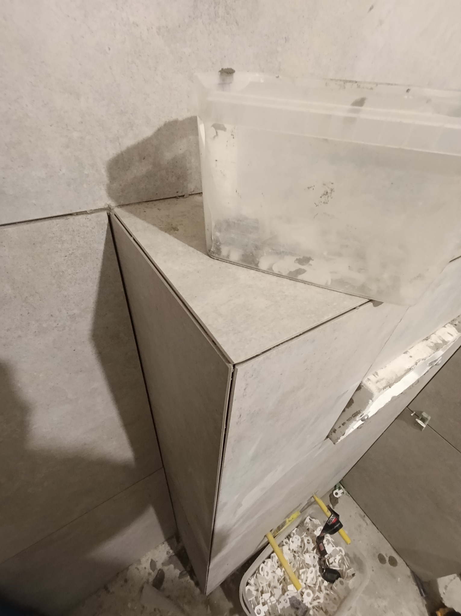 Układanie szarych płytek w narożniku pomieszczenia w Donimierzu, widoczne plastikowe elementy do poziomowania płytek i pojemnik z klejem.